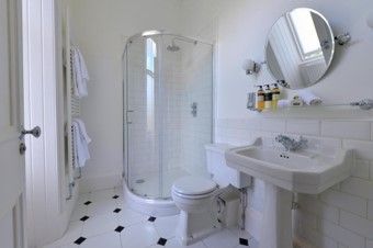 Bright white bathroom with glass shower, pedestal sink, round mirror, and black-and-white tiled floor