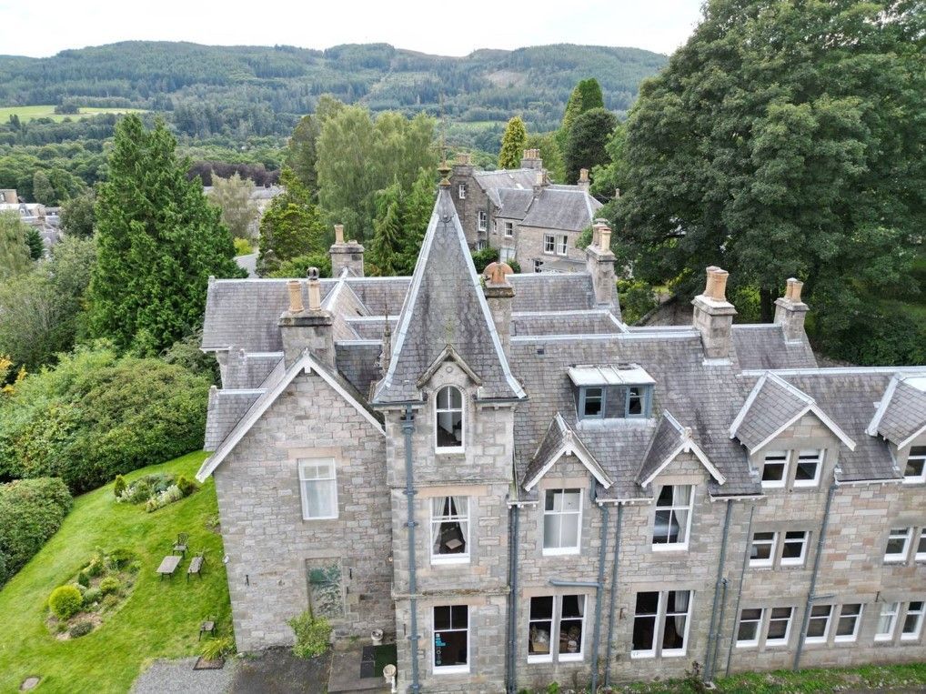 Stone mansion with steep gray roofs nestled among trees in a green hillside landscape