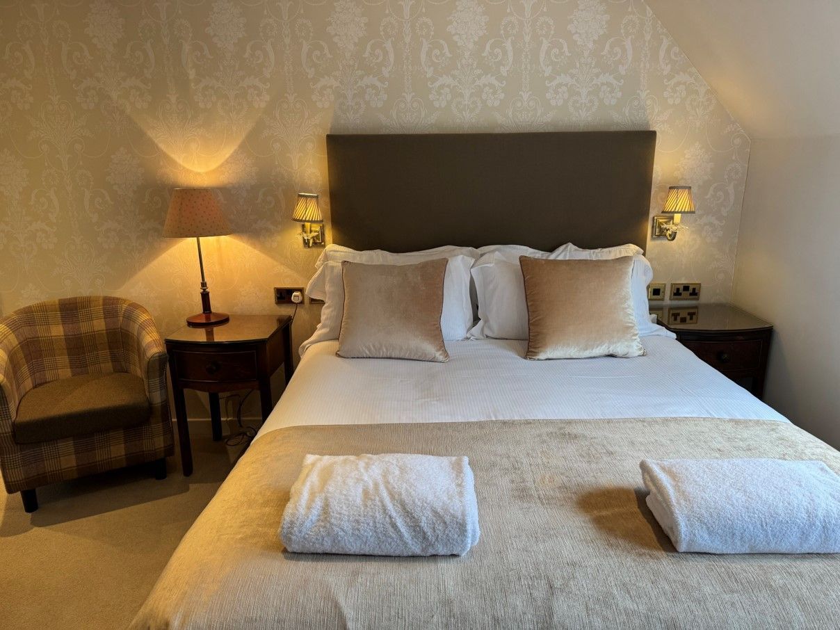 Neatly made bed with white linens and towels in a softly lit bedroom with bedside lamps and patterned wallpaper