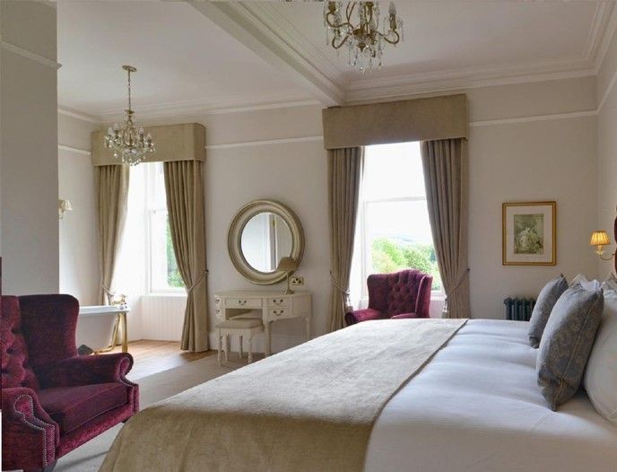 Elegant bedroom with a large bed, purple armchairs, a vanity mirror, and tall windows with drapes.