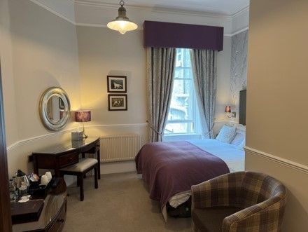 Cozy bedroom with a bed, window, desk, mirror, and plaid chair in soft purple tones