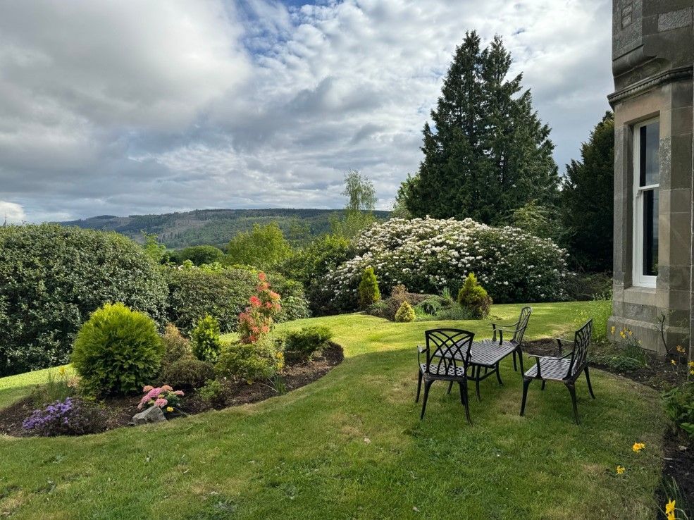 Garden patio with black chairs, lush lawn, flowering shrubs, and cloudy countryside views