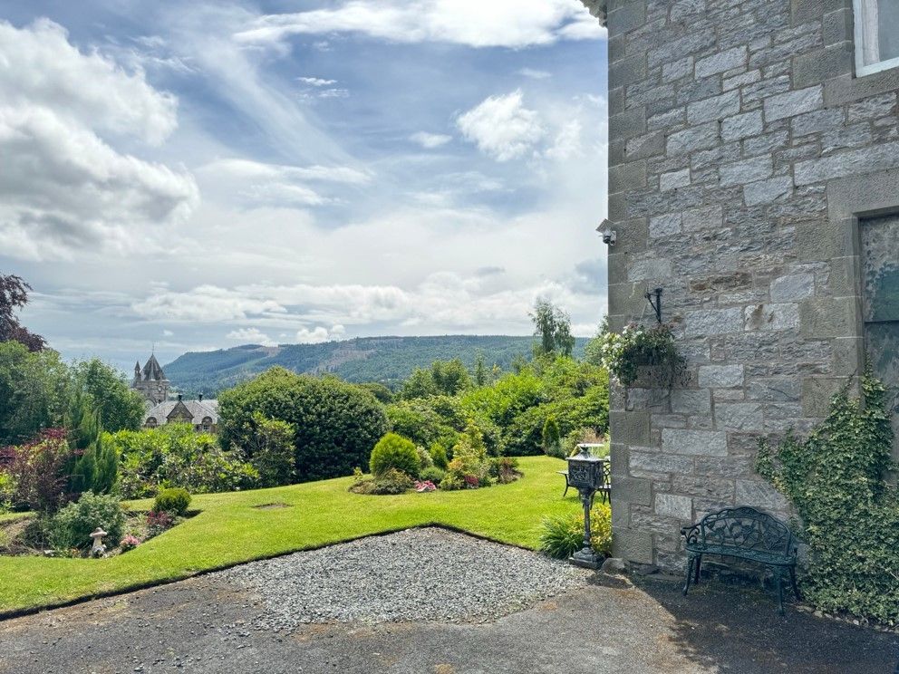 Stone house overlooking a green garden and gravel driveway, with mountains and cloudy sky in the distance