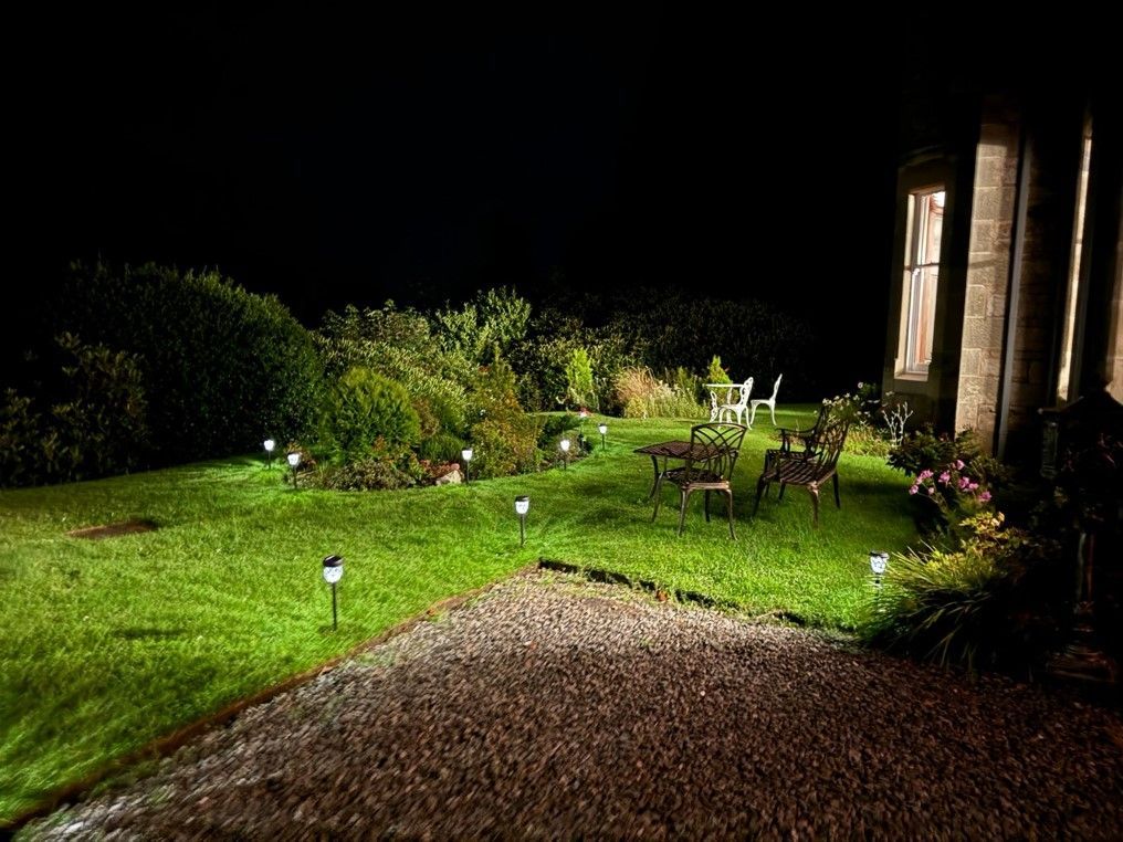 Nighttime garden path with lit lawn lamps, patio chairs, and a house exterior on the right