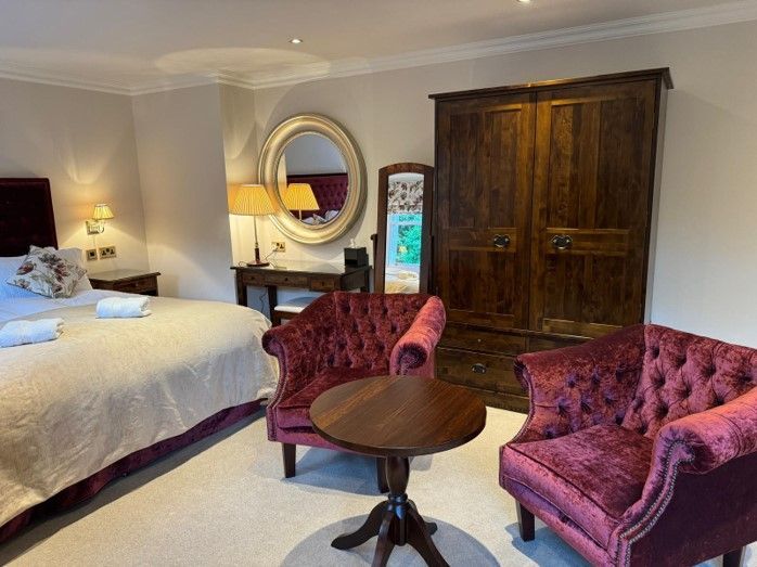 Bedroom with bed, burgundy chairs, round table, armoire, and mirror on beige walls