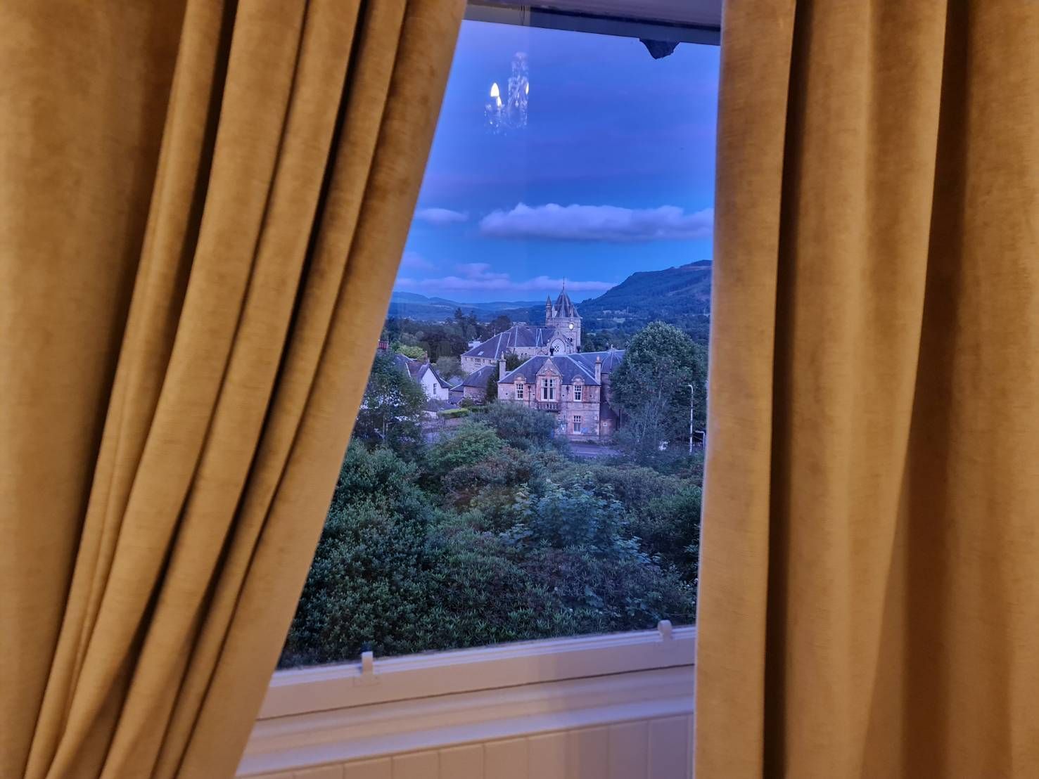 Curtained window overlooking a town and hills at dusk with blue sky
