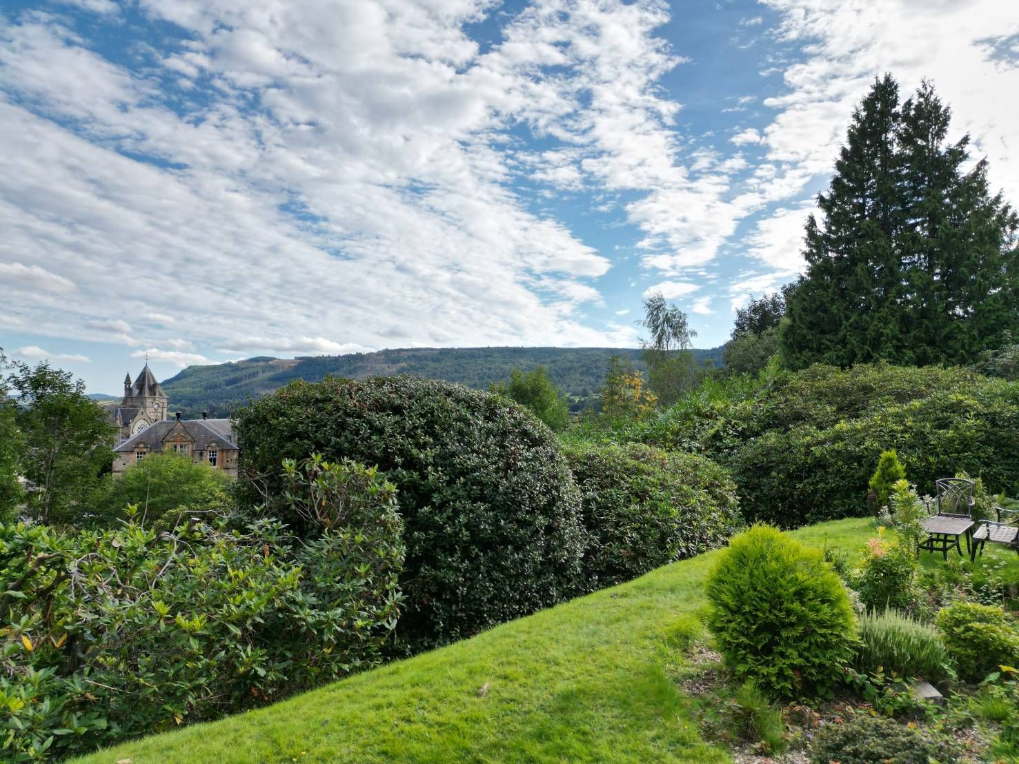 Lush garden hillside with trimmed hedges, trees, and a cloudy blue sky over distant hills