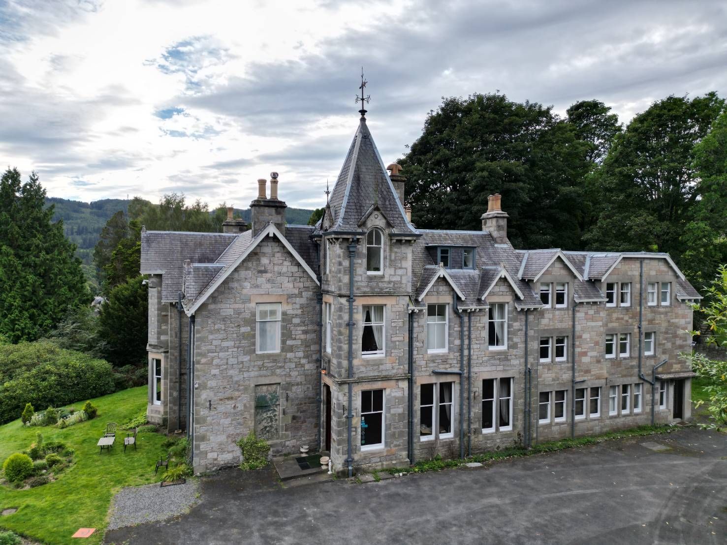 Stone manor house with a turret, set in a grassy hillside with trees and cloudy sky
