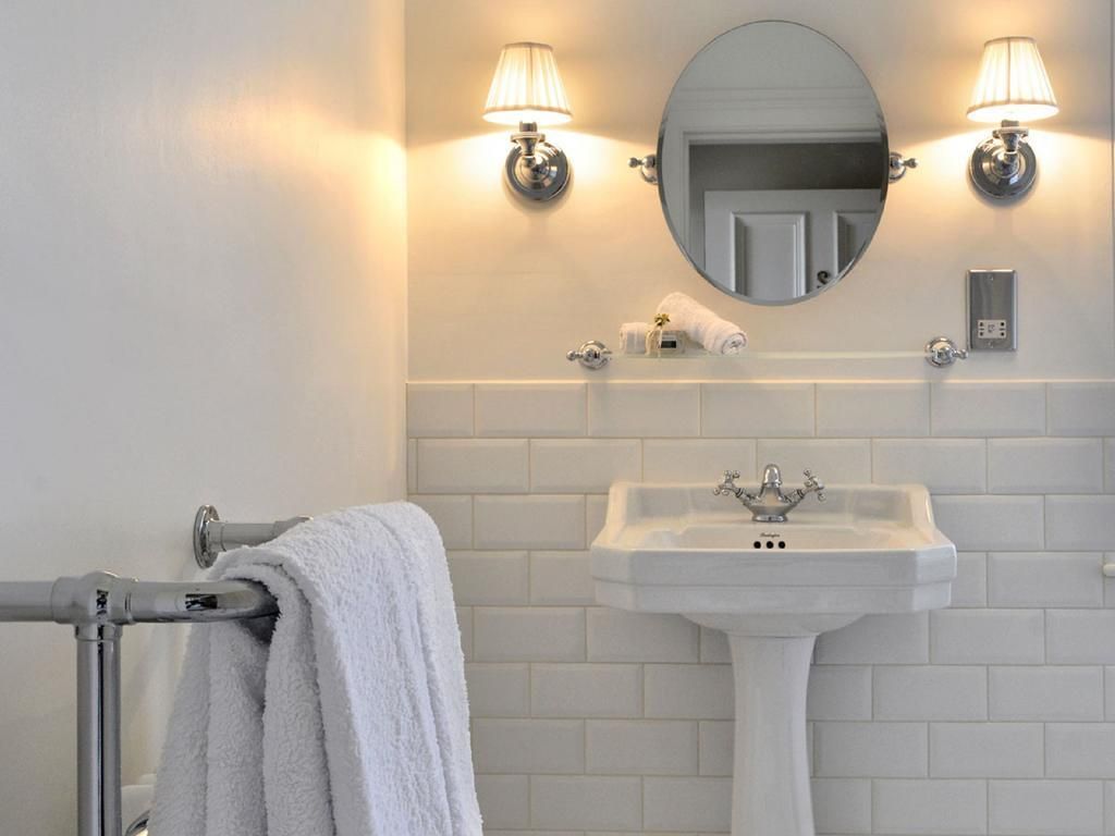 Bright white bathroom with pedestal sink, round mirror, sconces, and towel rack in the foreground.