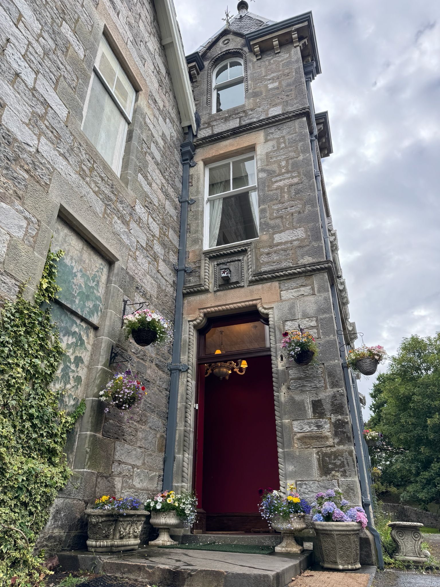 Stone building entrance with red door, hanging flowers, and cloudy sky above narrow courtyard