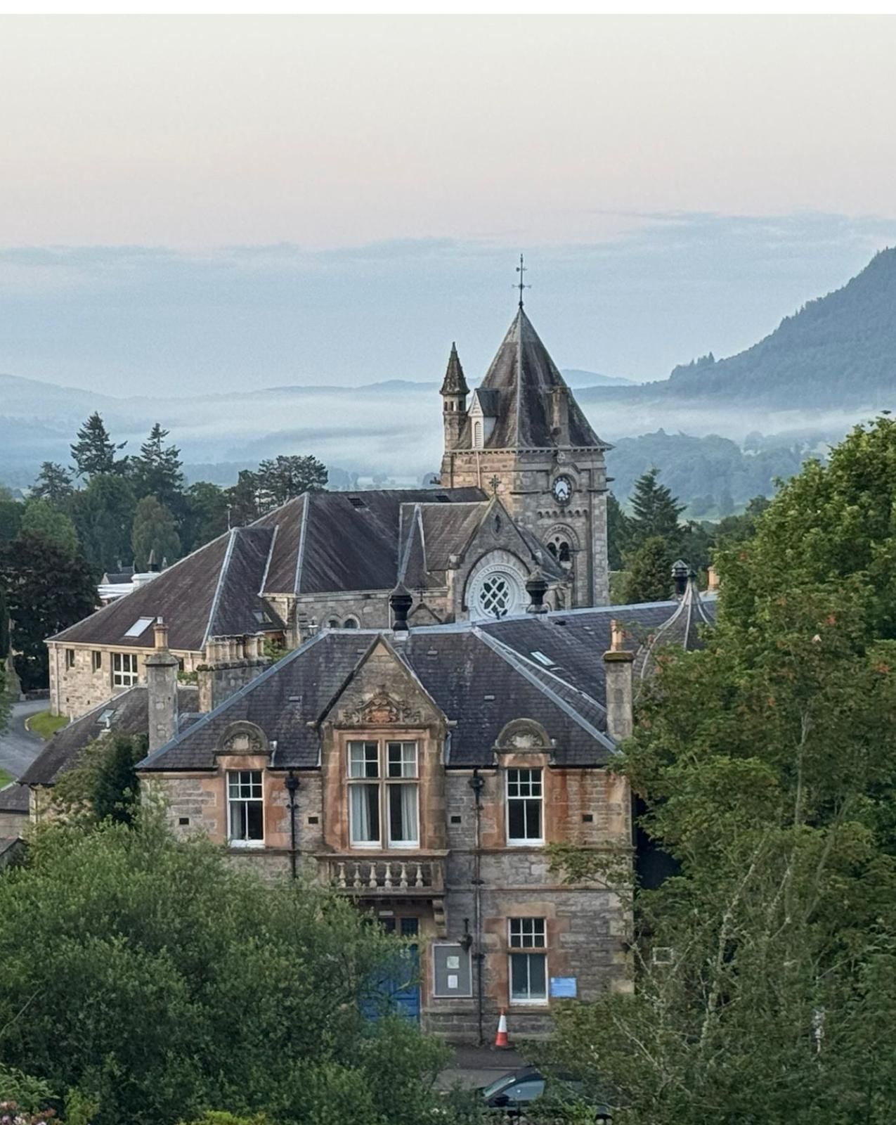 Stone manor with a tower amid trees, overlooking misty hills and a lake under a pale sky.