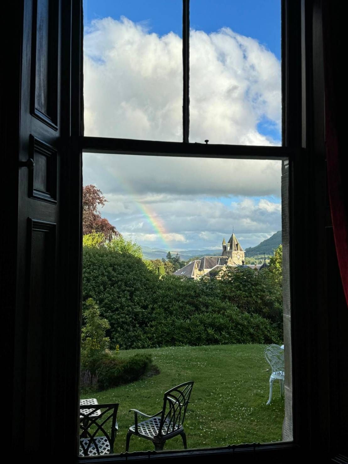 View through a window to a garden, chairs on grass, and a distant church under a cloudy sky