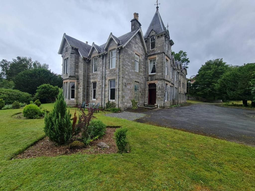 Stone manor house with steep gables, set beside a lawn and driveway under cloudy skies
