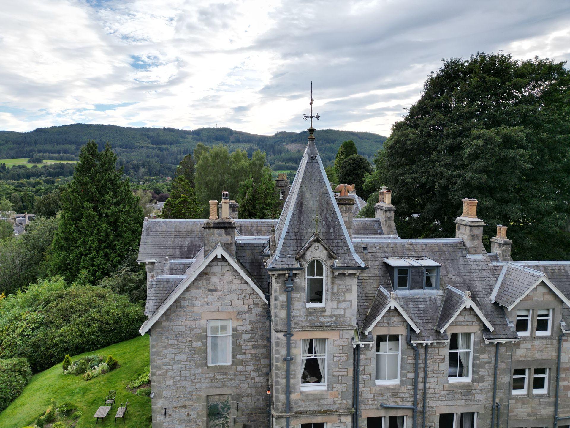 Stone manor house with slate roofs beside green hills under a cloudy sky