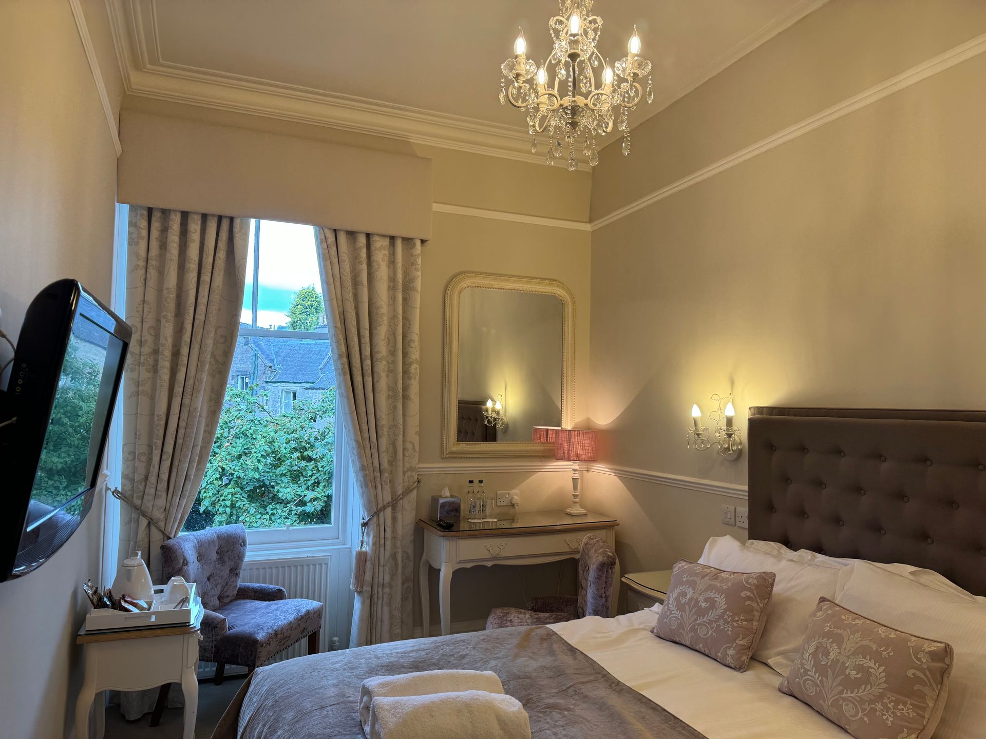 Elegant beige bedroom with a tufted bed, chandelier, vanity mirror, and window with curtains.