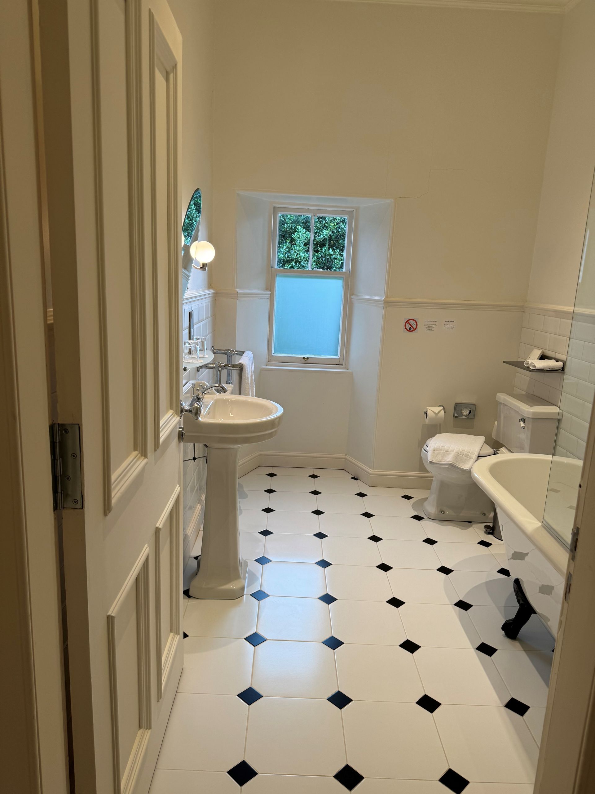 Bright bathroom with black-and-white tile, pedestal sink, toilet, and frosted window with greenery outside