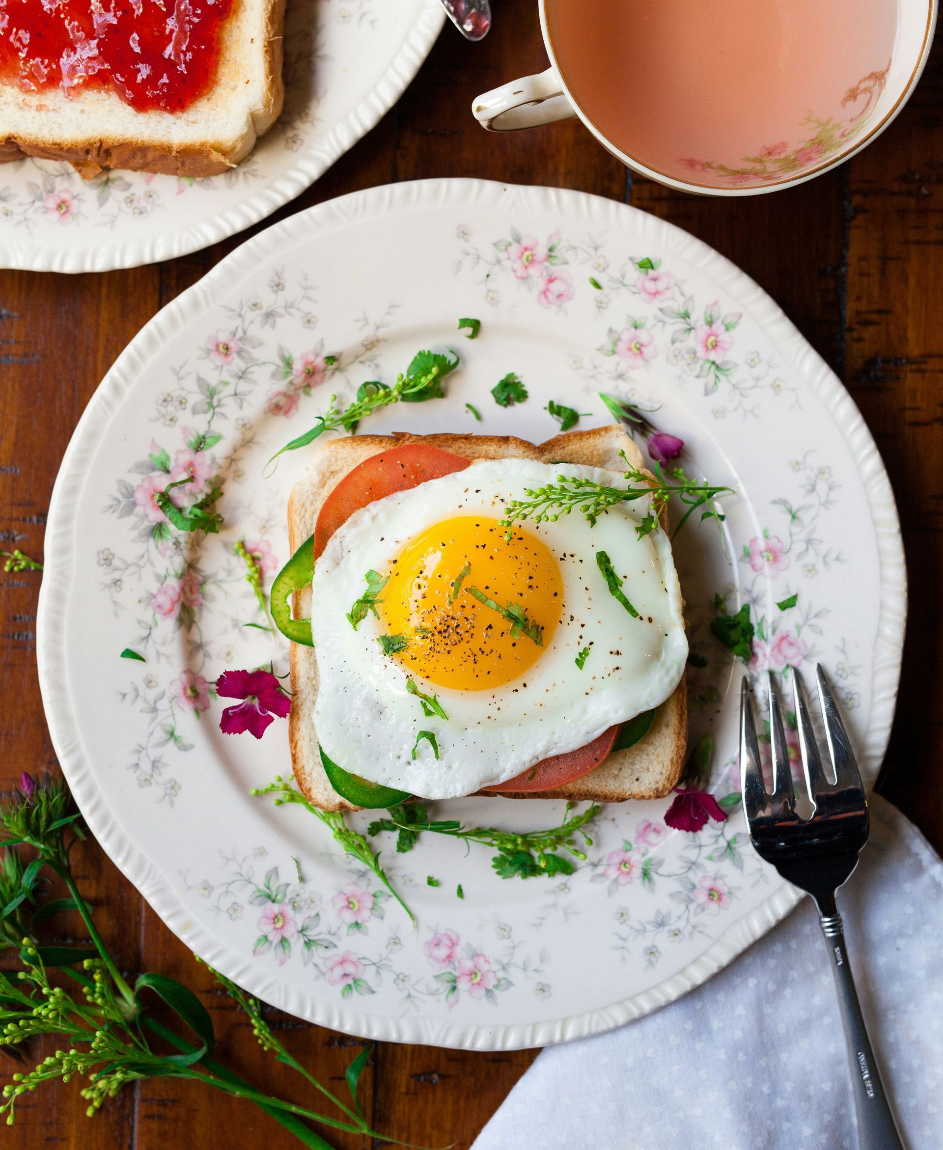 Open-faced egg sandwich on floral plate with tea and jam toast on a wooden table