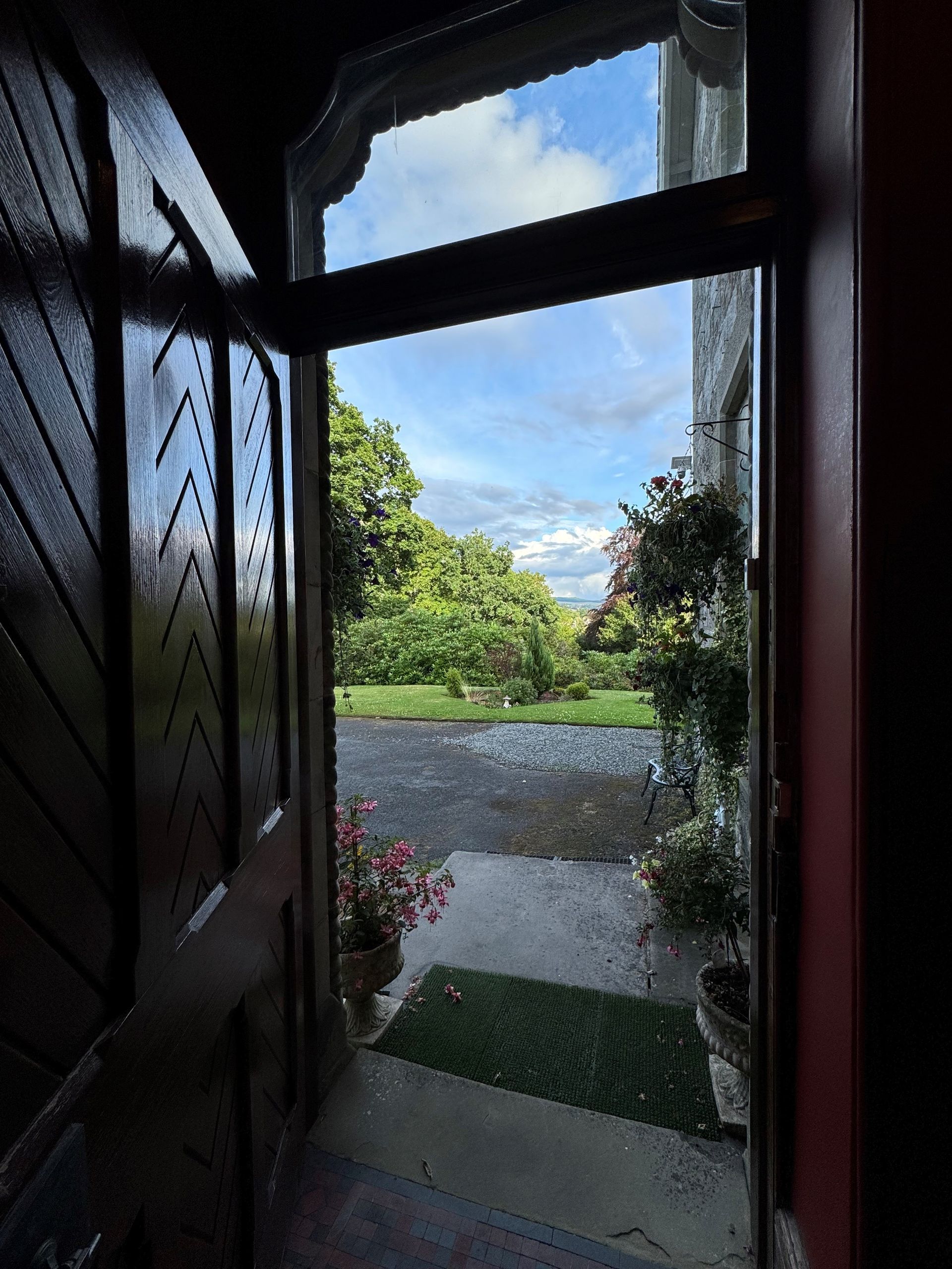 View from an open doorway to a garden path, green lawn, and blue sky with clouds.