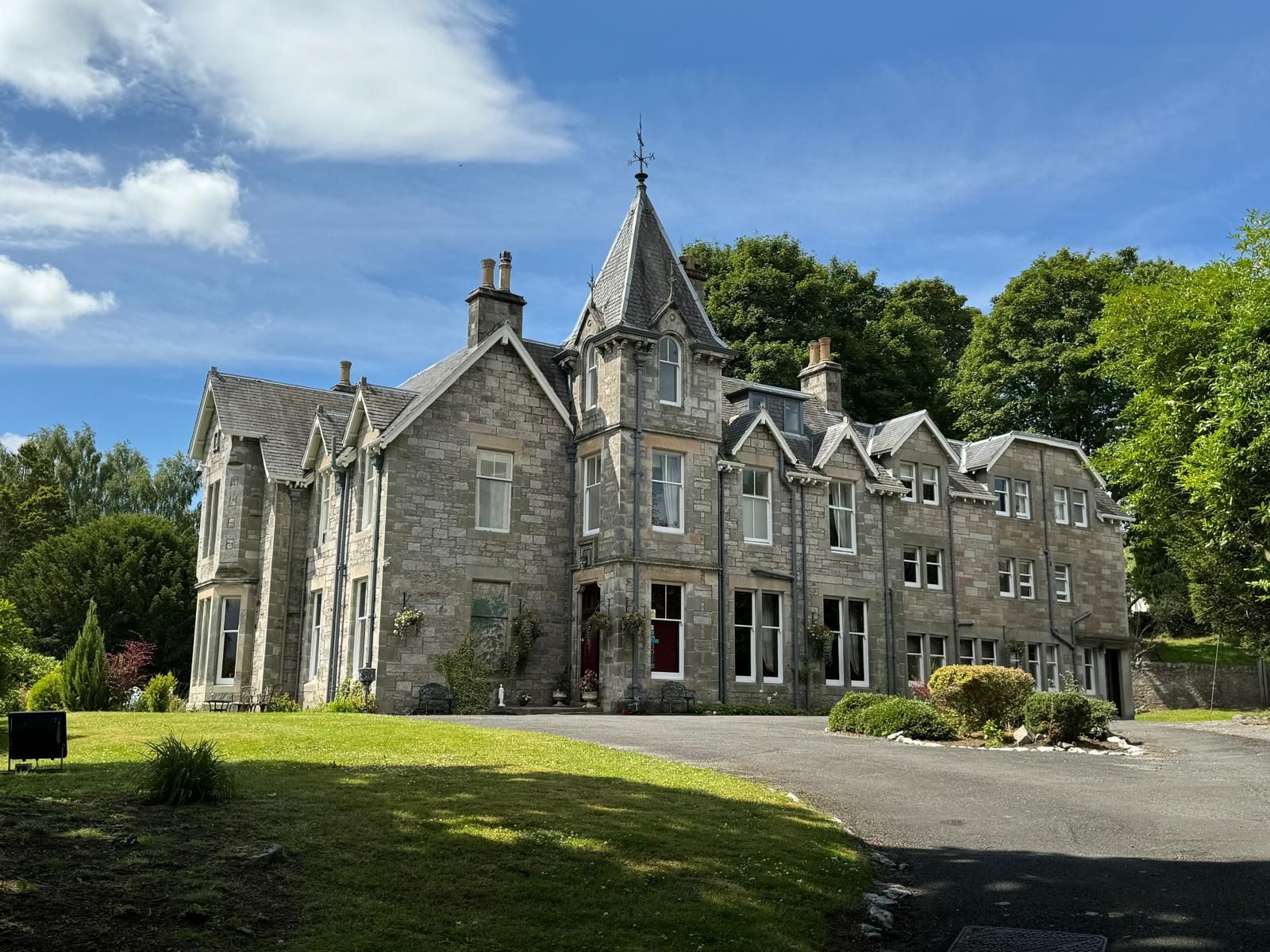 Stone manor house with a tall turret, set in a grassy landscaped garden under a blue sky.