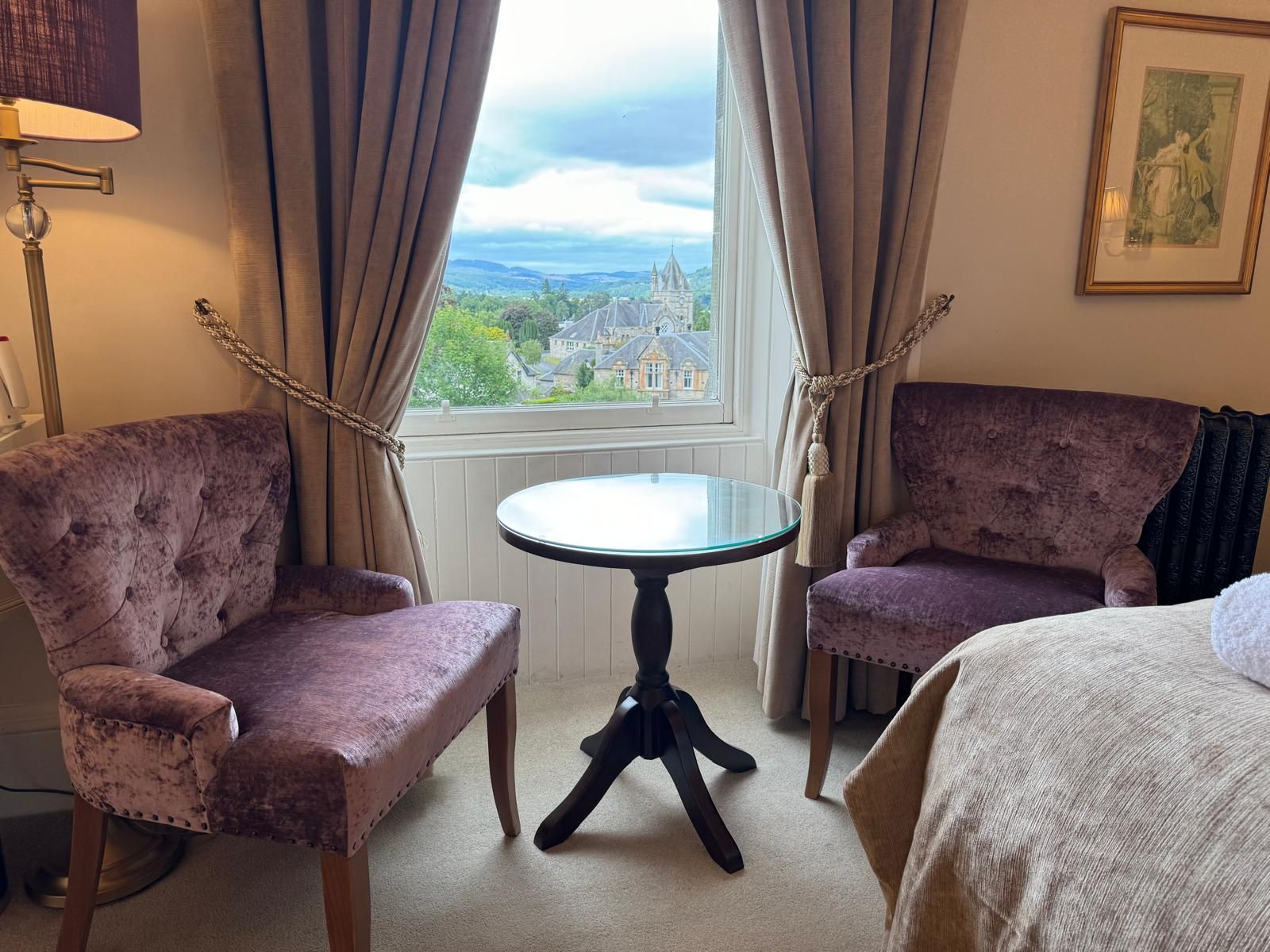 Cozy sitting room with two mauve chairs, round table, lamp, and a window view of greenery and hills