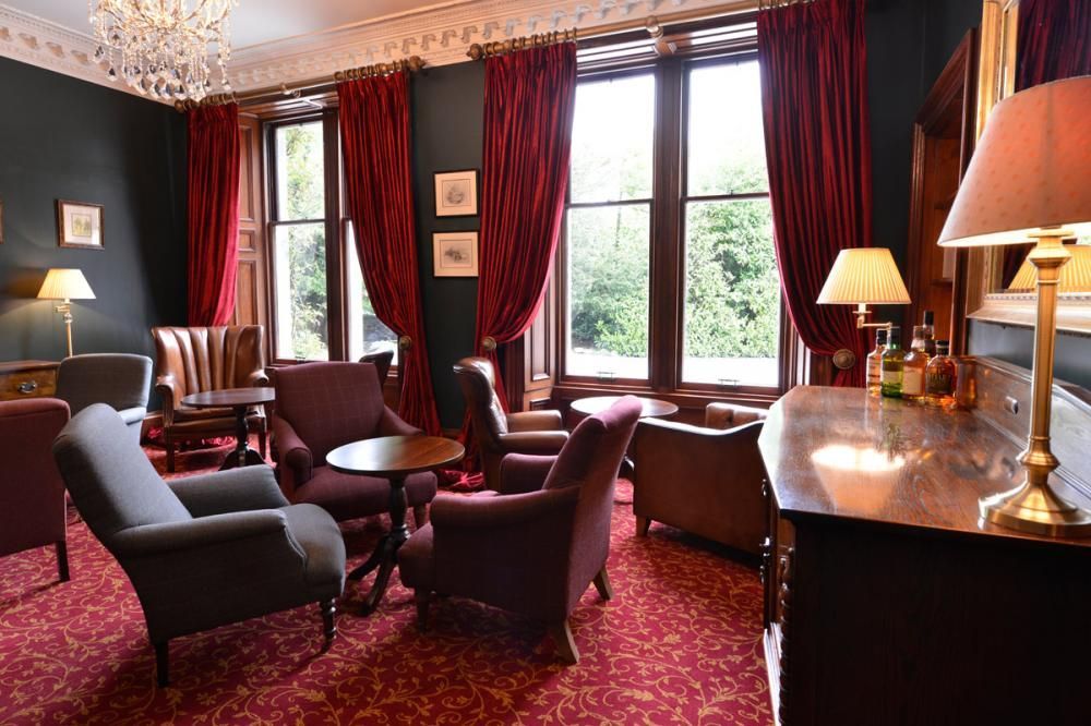 Elegant lounge with burgundy chairs, red carpet, tall windows, and a lamp on a dark wood sideboard