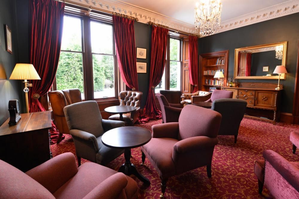 Elegant lounge with red carpet, pink chairs, wood desk, chandelier, and large windows with dark curtains
