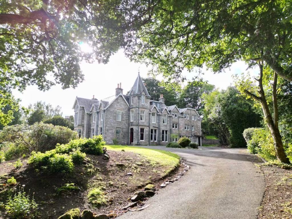 Stone manor house at the end of a winding driveway, framed by lush green trees and shrubs.