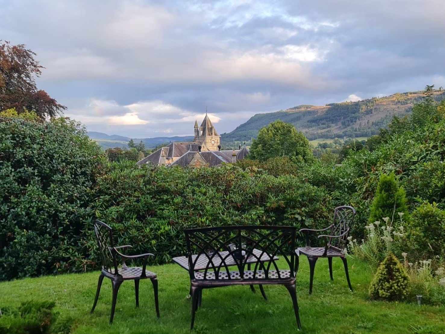Garden chairs on a lawn overlooking a distant castle and green hills under a cloudy sky