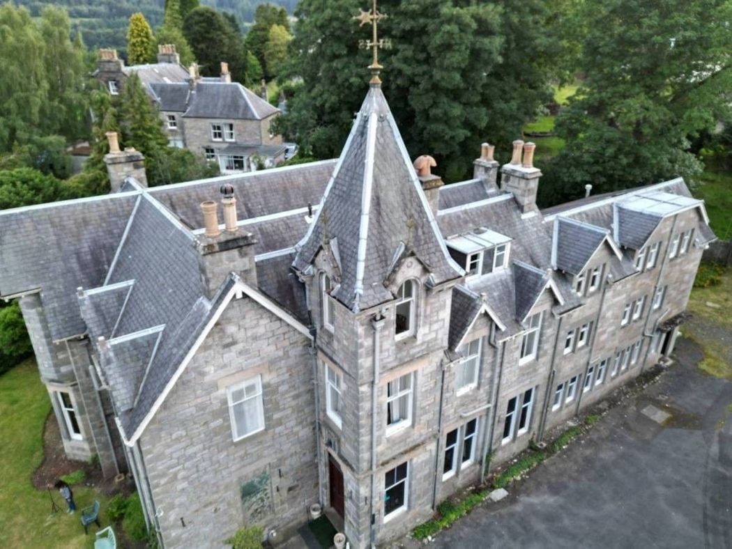 Aerial view of a gray stone manor with steep roofs and a central tower, surrounded by trees