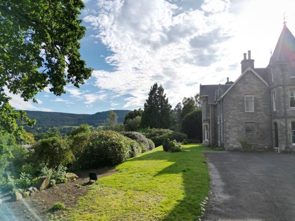 Stone house beside a sunny garden path with trees, hills, and a bright cloudy sky