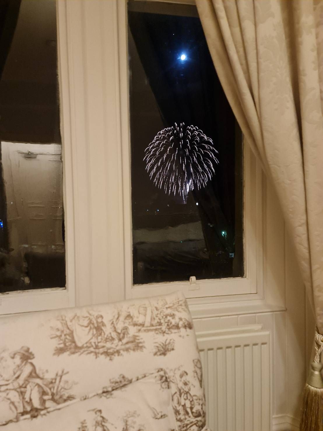 Window view of a night sky with fireworks above a radiator and patterned chair by curtains