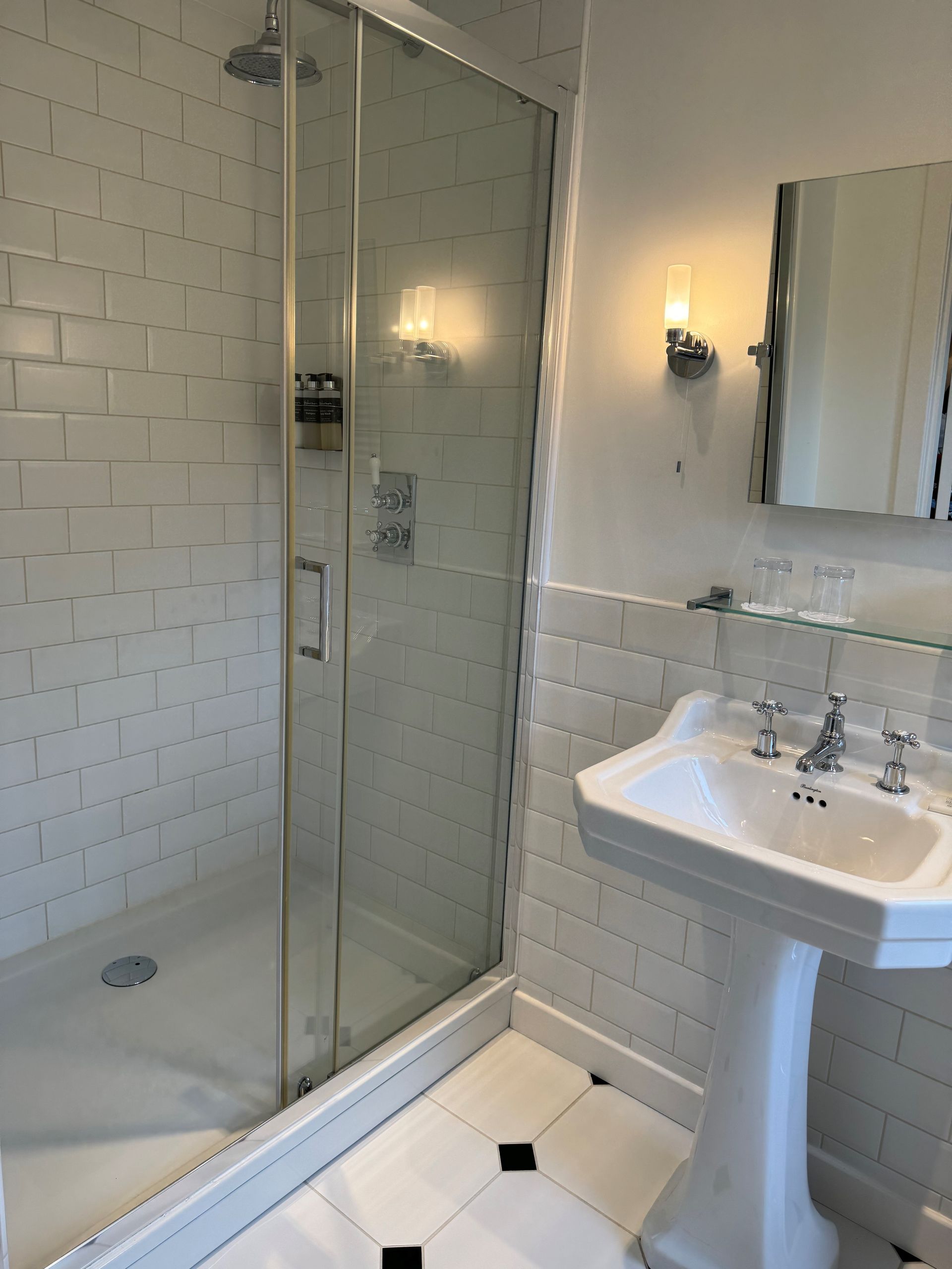 Small white-tiled bathroom with a glass shower, pedestal sink, and wall mirror.