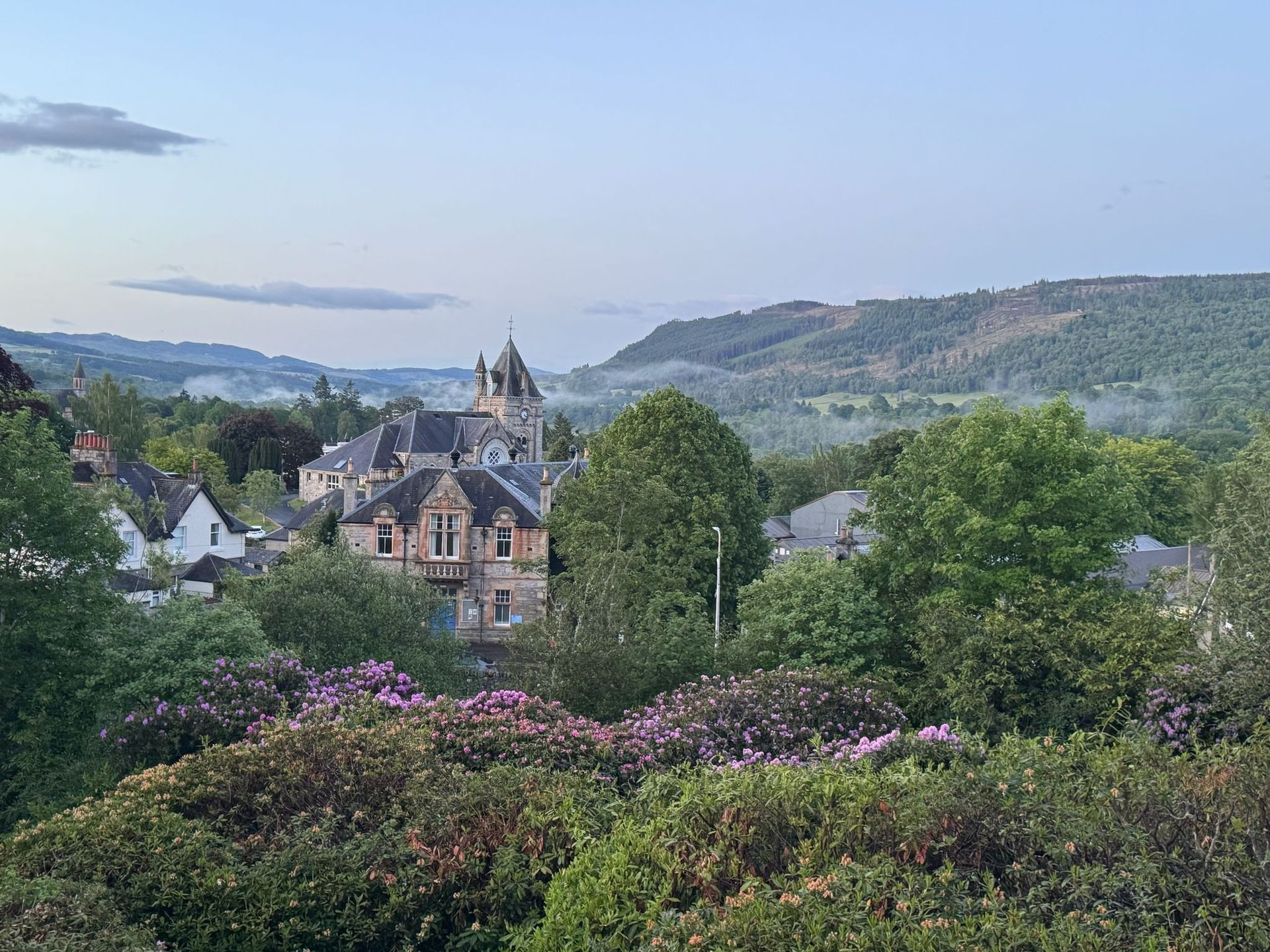 Historic stone house amid lush green trees and purple flowers, overlooking rolling hills under a pale blue sky