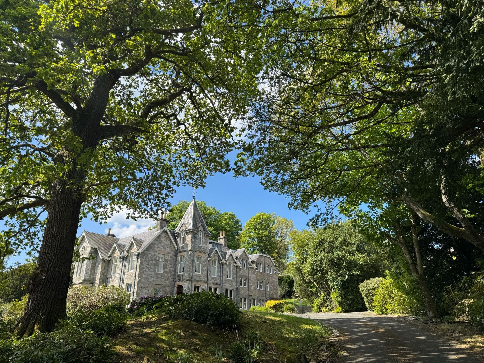 Stone manor house with spires beside a tree-lined road under a blue sky