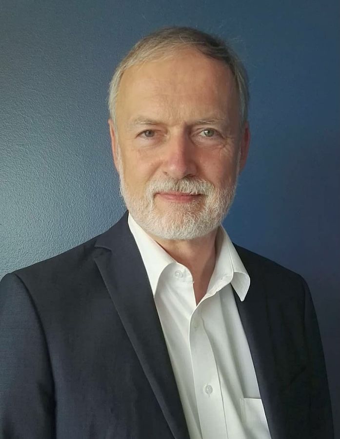 Michael Schack en costume sombre et chemise blanche, souriant, sur fond bleu.