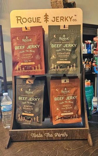 Jerky display stand with four Rogue Jerky packs. Brown, green, red, and black packaging, wooden stand, text