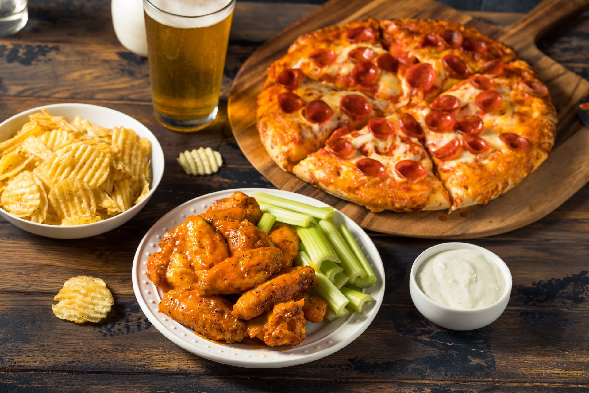 A pizza , wings , chips and a glass of beer on a wooden table.