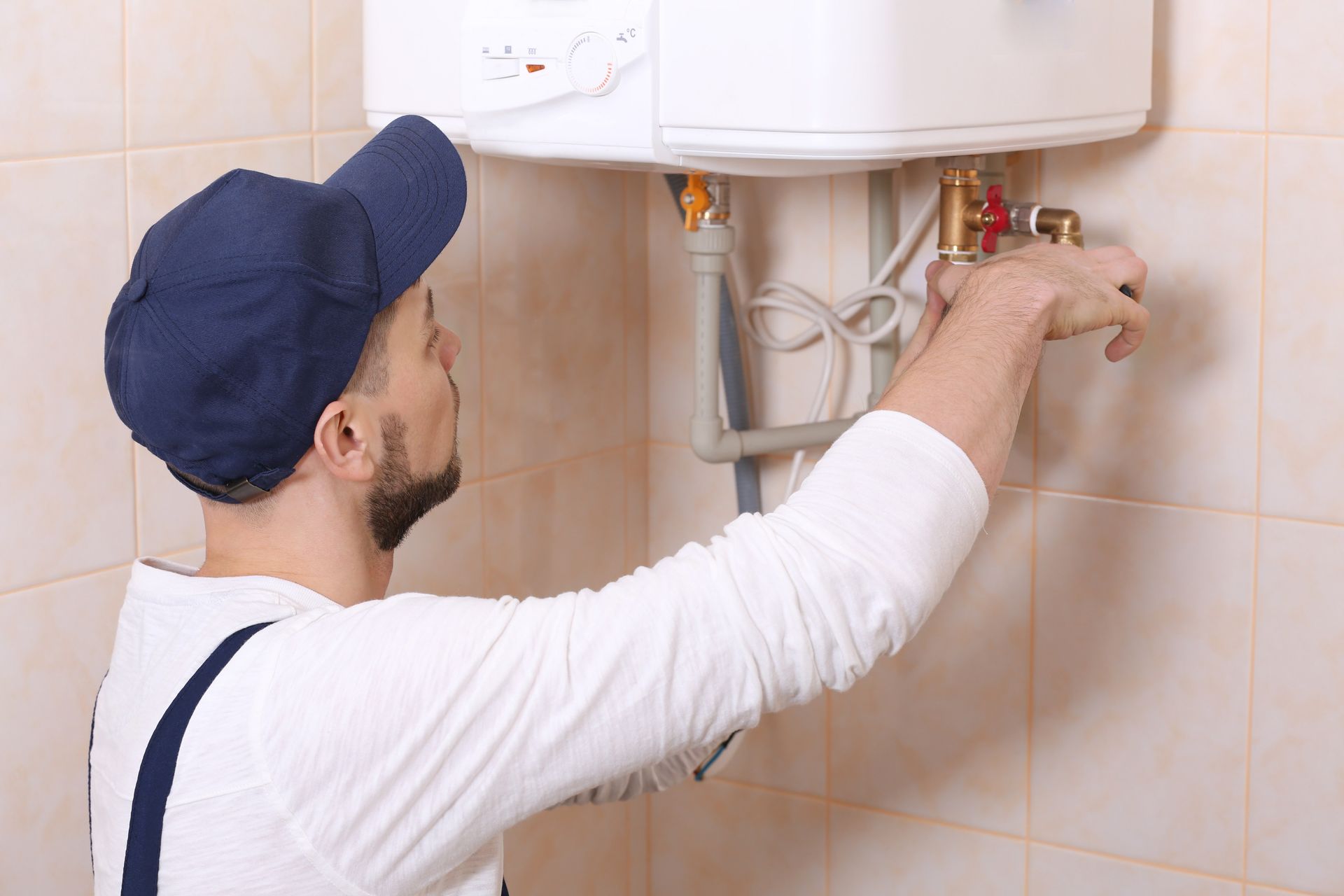 Plumber adjusting pipes under a white wall-mounted water heater in a tiled bathroom