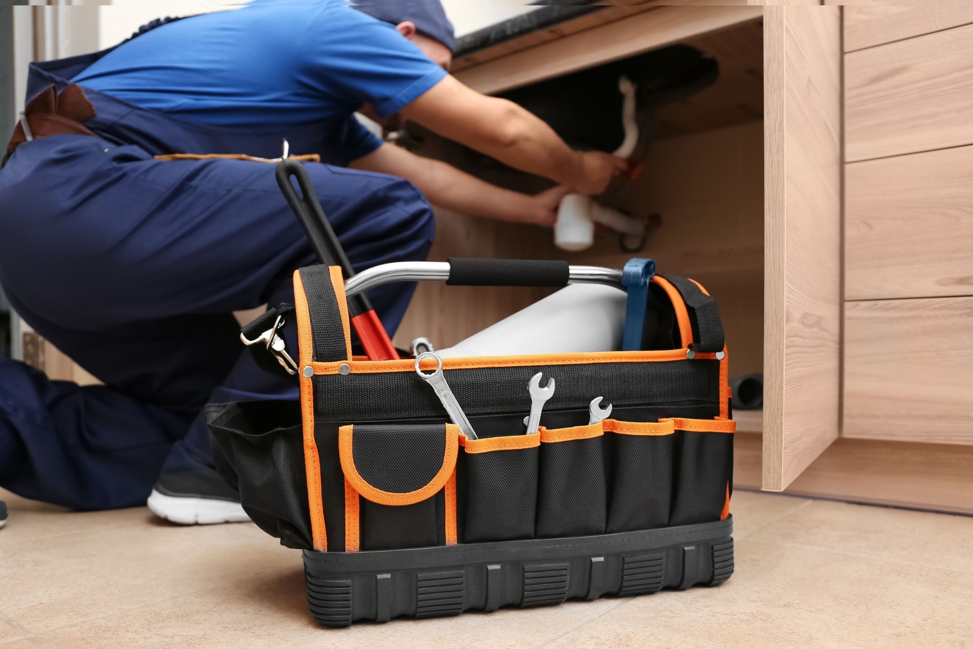 Technician kneeling under a cabinet with a black-and-orange tool bag in the foreground.