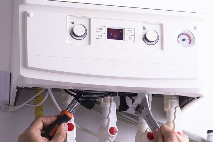 Hands repairing a white wall-mounted boiler with tools underneath.