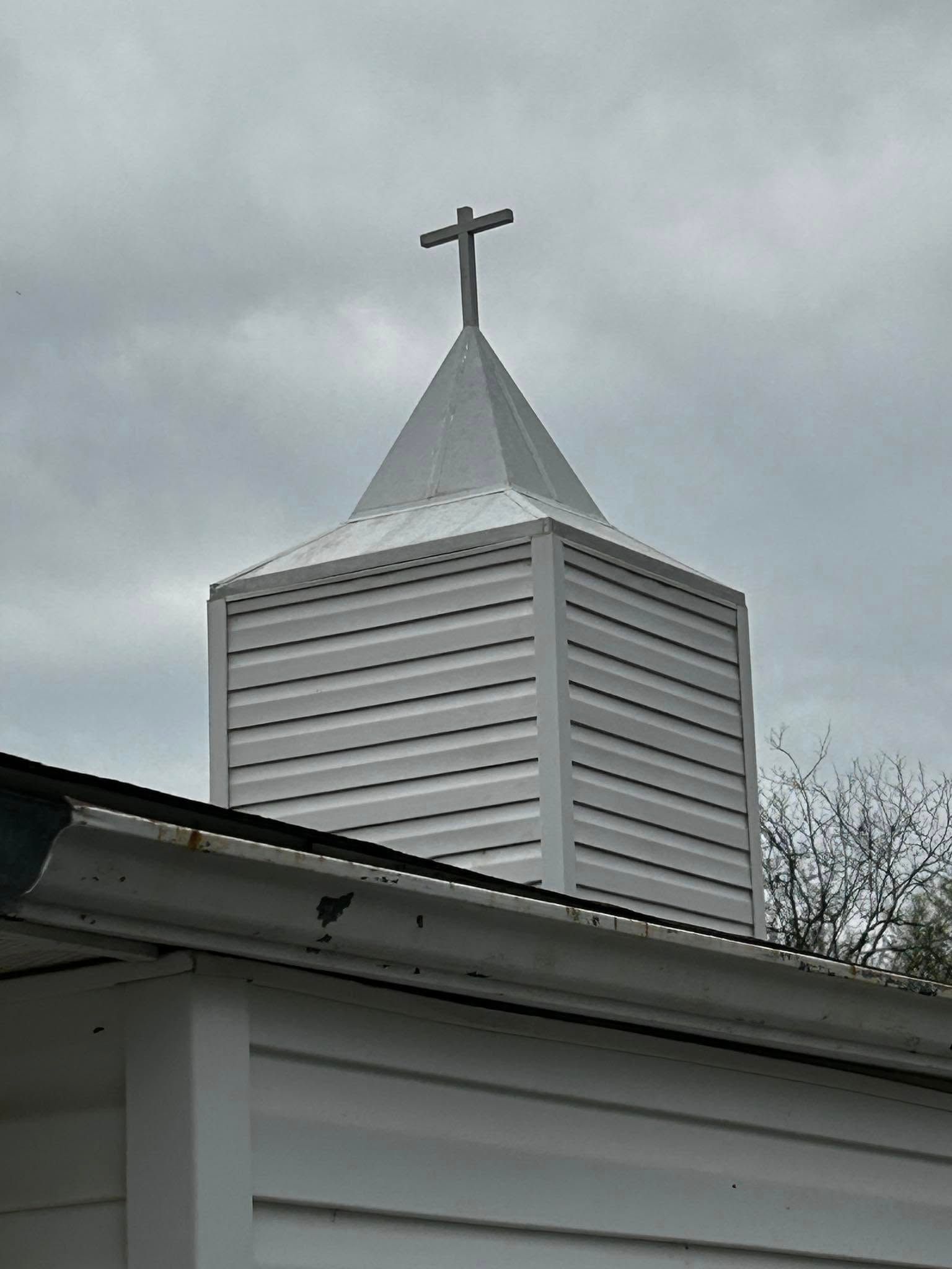 White church steeple with a cross above a building roof under a cloudy sky
