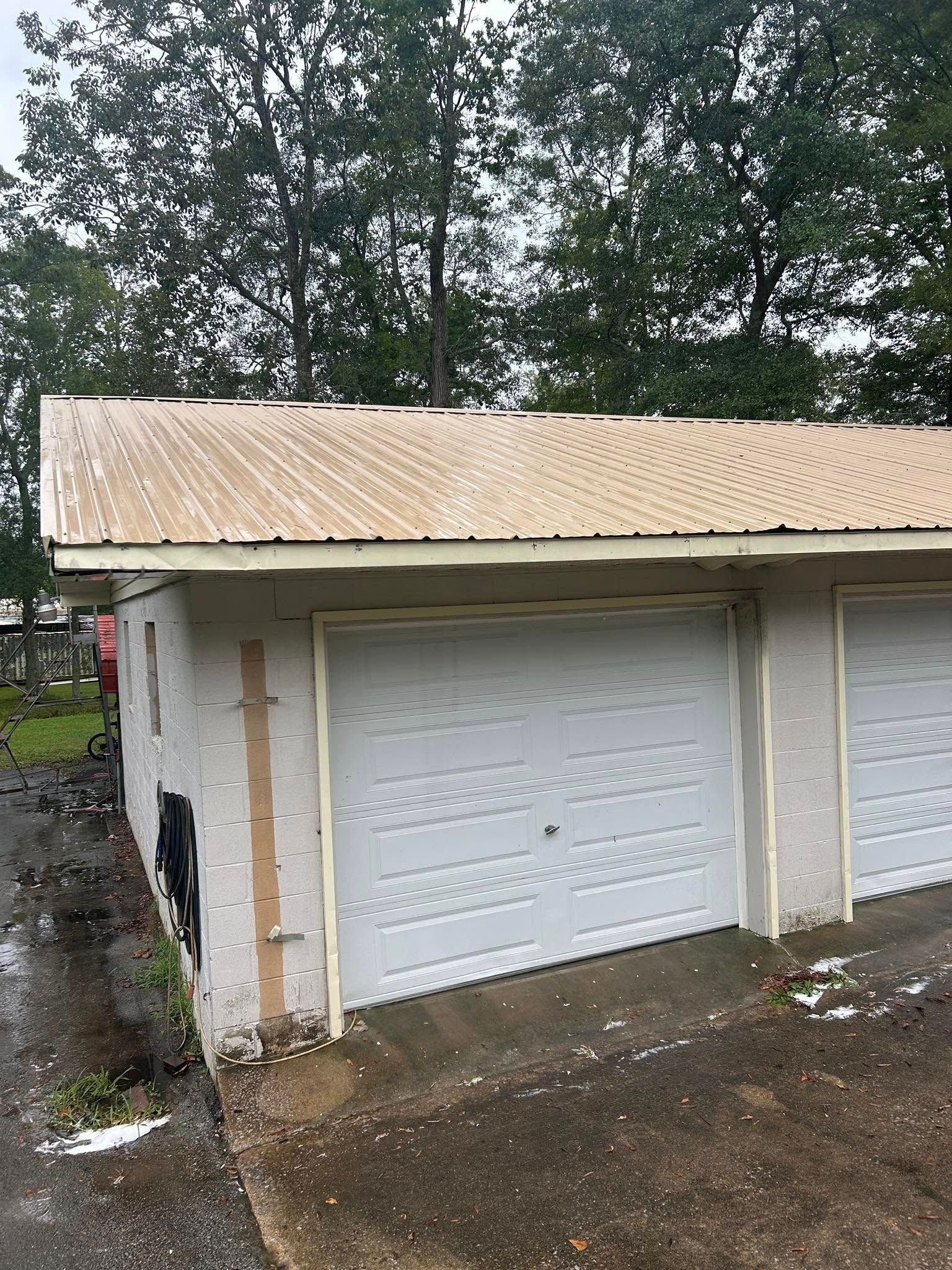 White garage with tan roof and trim, front view beside a wet driveway and trees.
