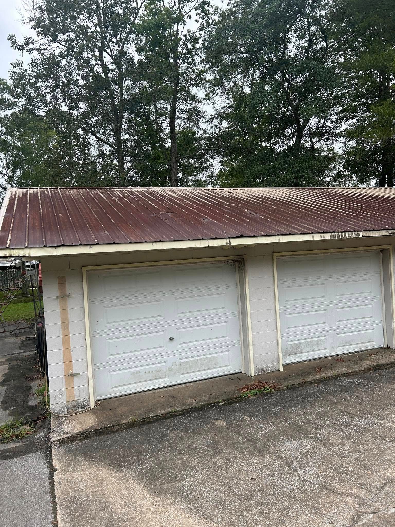 Two white garage doors beneath a rusted metal roof, with trees in the background.