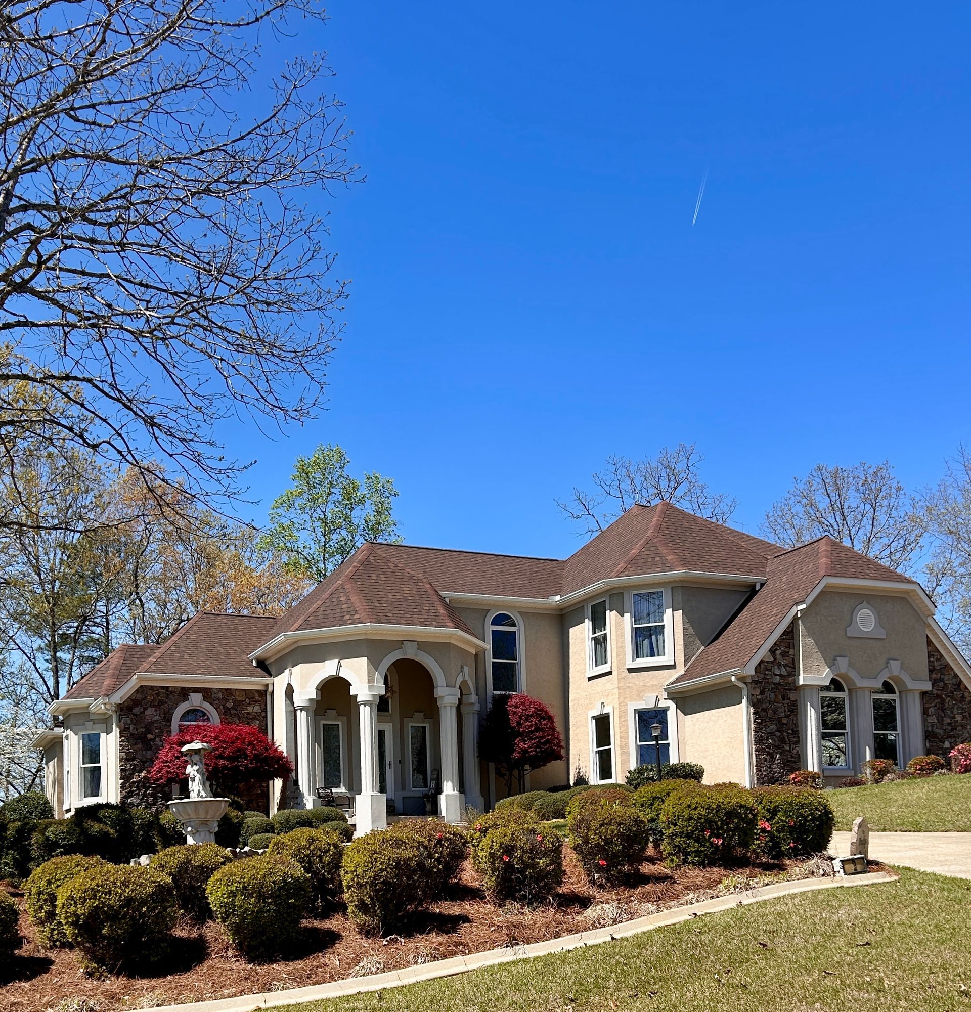 Large beige house with a brown roof, white columns, and manicured shrubs under a clear blue sky