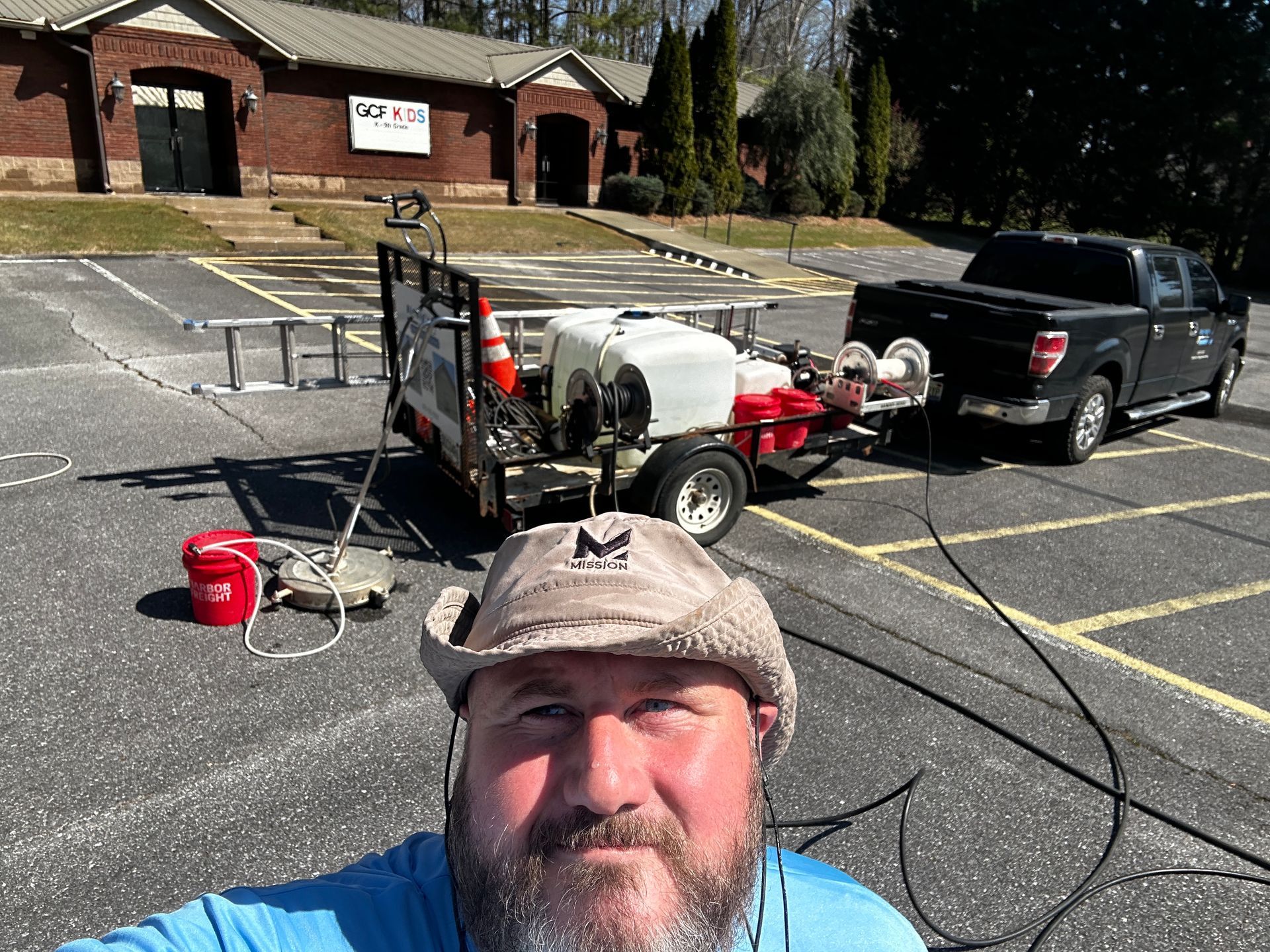 Man taking selfie beside a utility trailer and equipment in a parking lot outside brick buildings