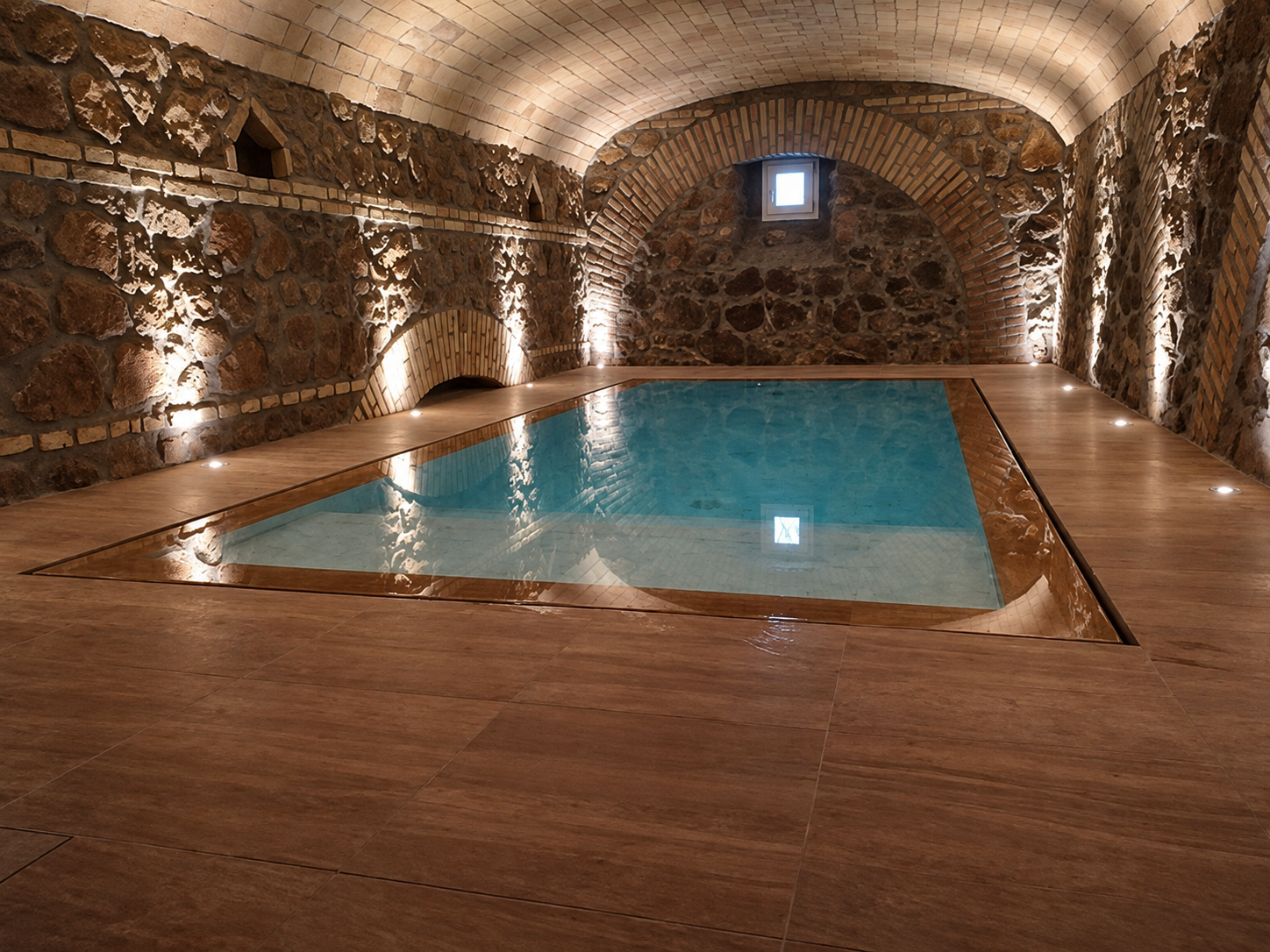 Piscina coperta con pareti in pietra, soffitto a volta e pavimento in legno, illuminata da una piccola finestra.