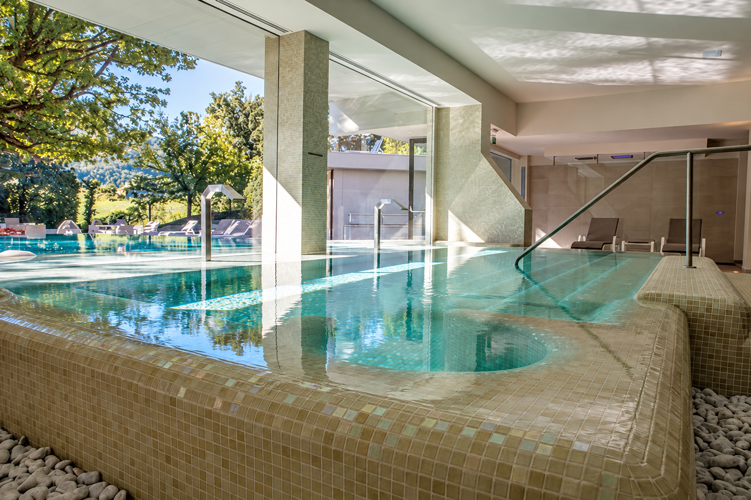 Piscina termale coperta con acqua limpida, bordo in mosaico e vegetazione all’esterno visibile attraverso le grandi finestre