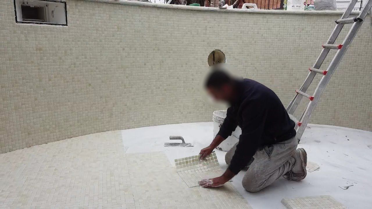 Persona inginocchiata sul bordo bianco della piscina, al lavoro vicino a pareti piastrellate, con una scala sullo sfondo