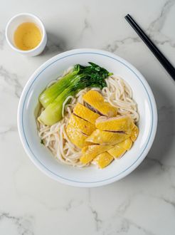 Bowl of noodles topped with sliced chicken and bok choy, with dipping sauce and chopsticks on a marble table