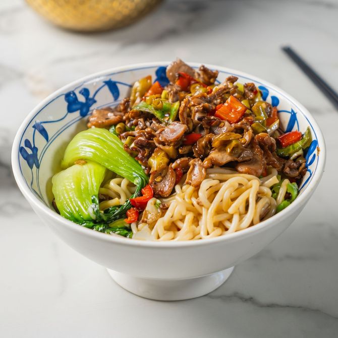Bowl of noodles topped with stir-fried beef and vegetables, with bok choy on a marble table